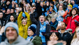 Crowd of Manchester City fans celebrating in a stadium, with visible team logos on clothing.