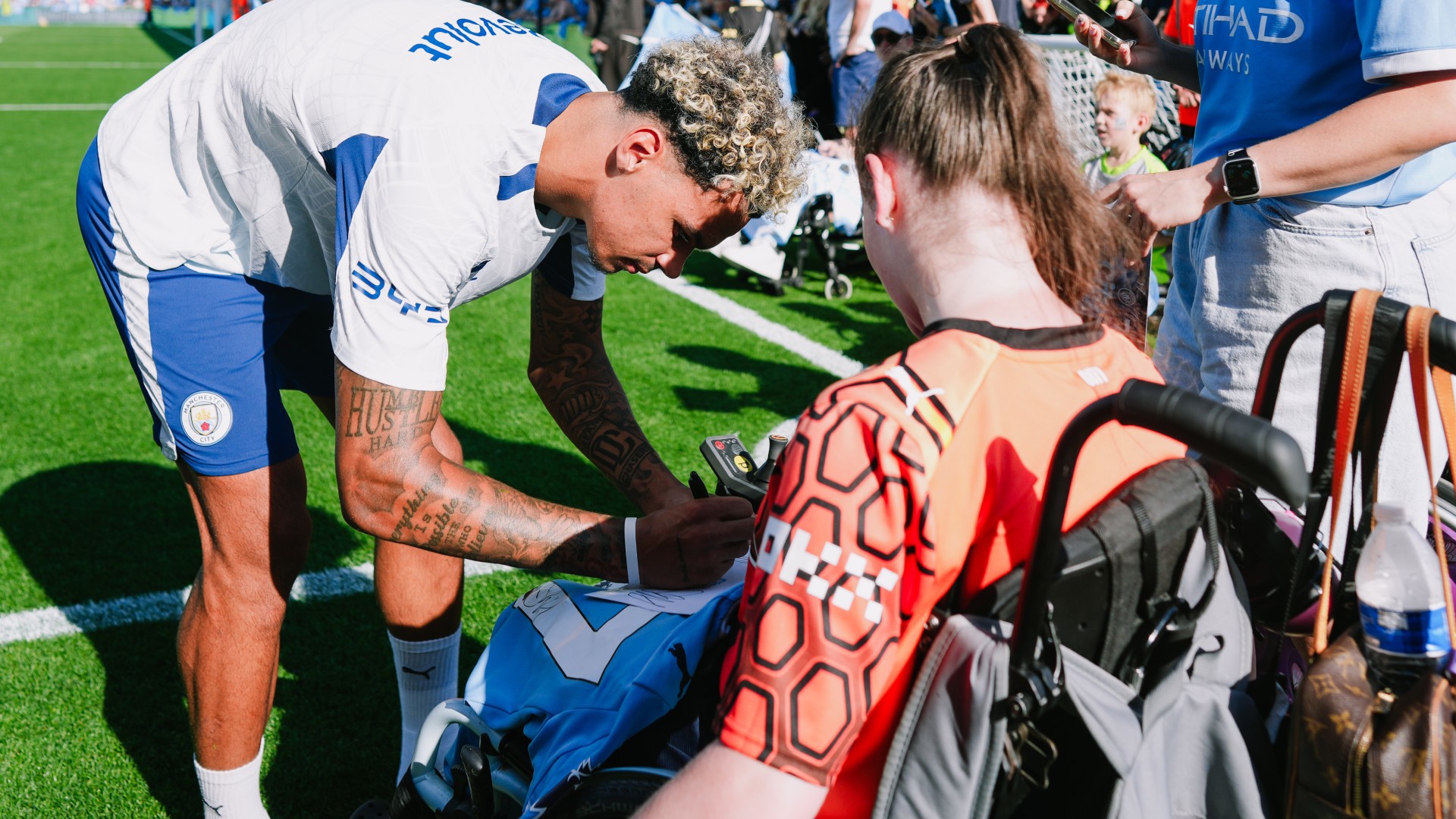 Manchester City player signing autographs for fans, including a fan in a wheelchair, on a soccer field.