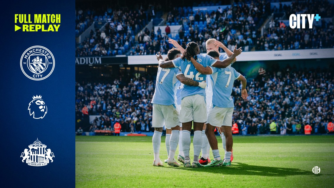 Manchester City players celebrate on the field after a goal, huddled together in light blue jerseys. The crowd is visible in the stadium background. Premier League and Sunderland logos are on the left side.
