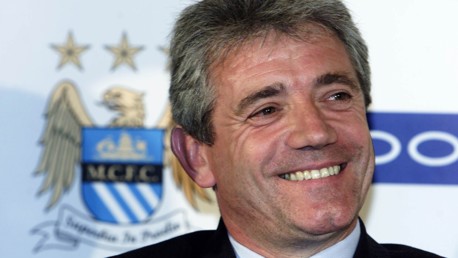A person smiling in front of a backdrop featuring the Manchester City Football Club crest.