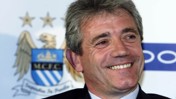 A person smiling in front of a backdrop featuring the Manchester City Football Club crest.