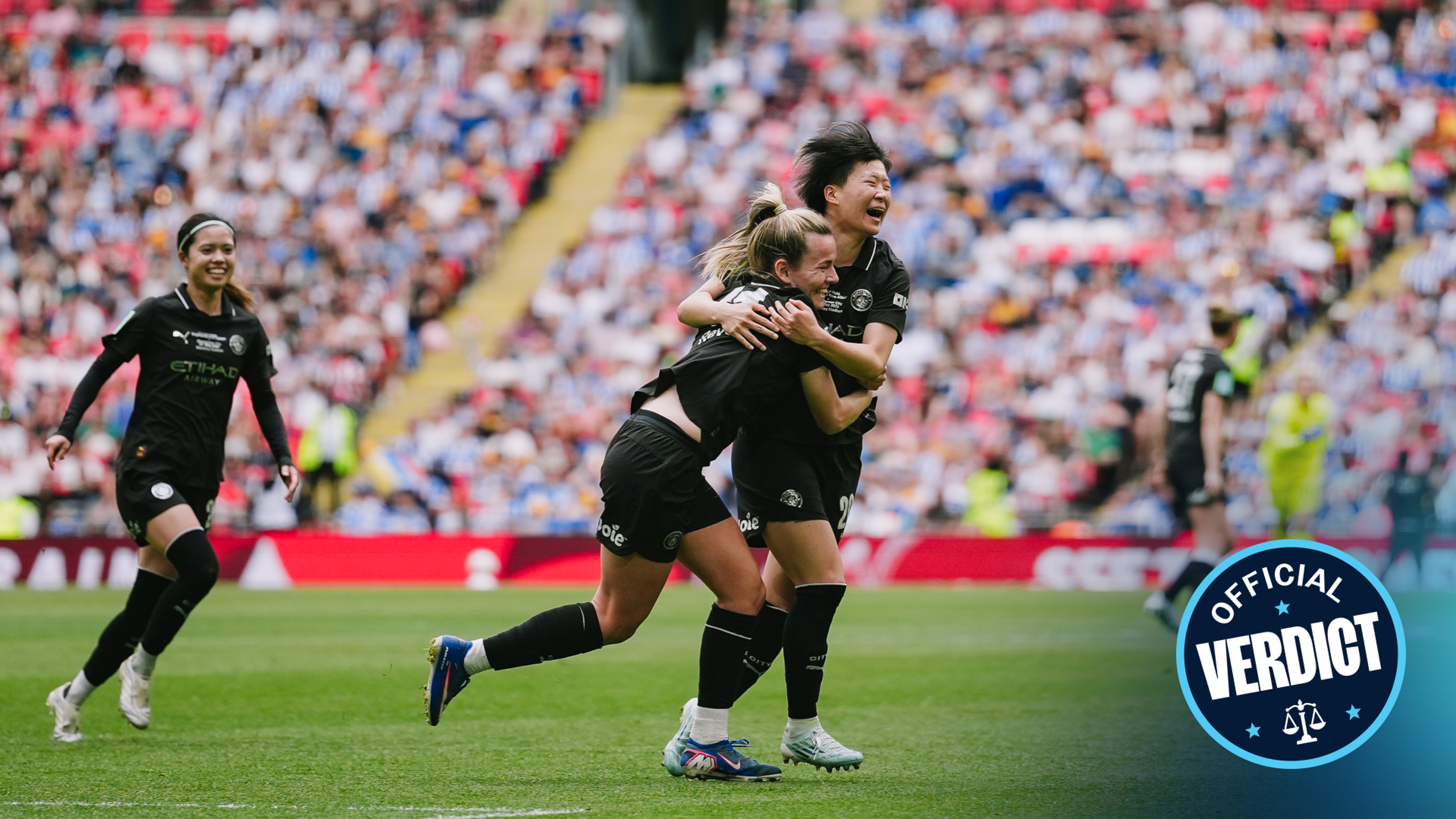 Women's football team celebrates a goal on a stadium with a crowd. Players wearing black jerseys and cheering enthusiastically.