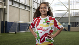 A young girl wearing a Kellogg's branded football jersey with colorful abstract patterns, standing on an indoor football pitch with hands on her hips.