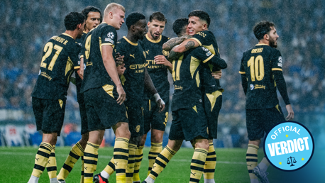 Manchester City players celebrating a goal under rain in a match, wearing dark jerseys with yellow accents. Some players are hugging while others are cheering.