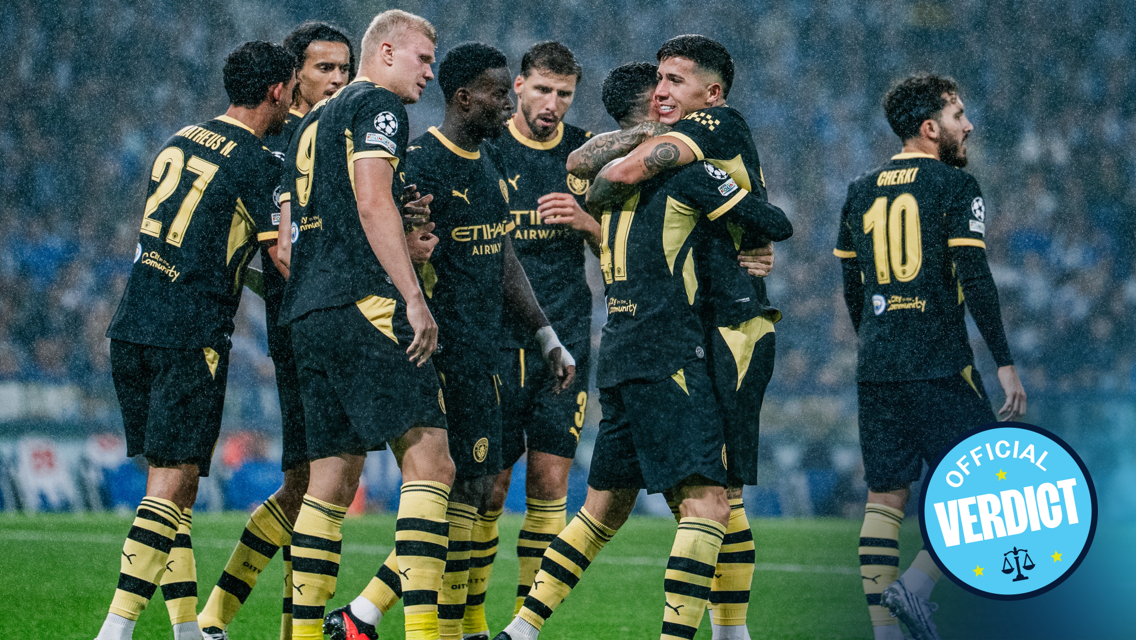 Manchester City players celebrating a goal under rain in a match, wearing dark jerseys with yellow accents. Some players are hugging while others are cheering.