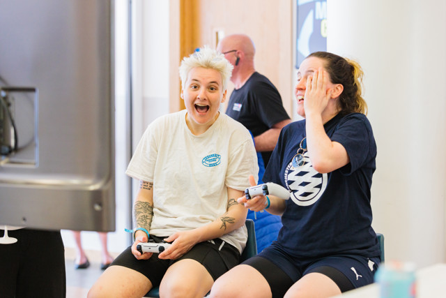 Two individuals are seated while playing video games with controllers. They appear focused on the screen.