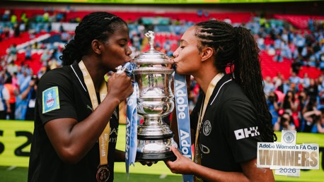 Two players celebrate by kissing the trophy after winning the Adobe Women's FA Cup in 2026. They are wearing black jerseys with gold medals around their necks.