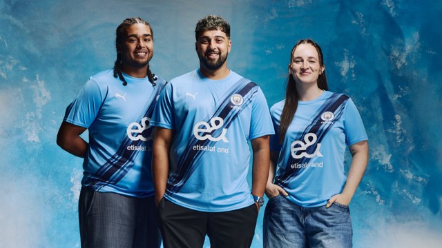 Three individuals wearing the 2023 Manchester City home kit featuring sponsor 'etisalat and' and the Puma logo, standing against a blue backdrop.