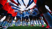 A group of soccer players in orange vests and blue uniforms gather in a huddle on a green field with their hands extended towards the center.