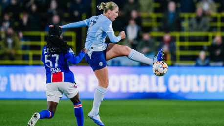 Two soccer players in action during a match. One player in a light blue kit attempts to control the ball mid-air while the opponent in a blue and red kit challenges for possession.
