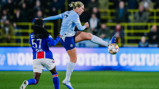 Two soccer players in action during a match. One player in a light blue kit attempts to control the ball mid-air while the opponent in a blue and red kit challenges for possession.