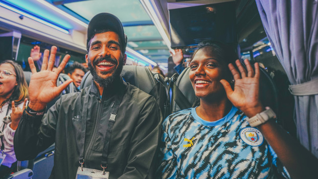 A group of fans are on a bus, with one person wearing a Manchester City jersey with wavy stripes and waving at the camera.