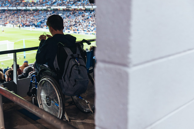 A person in a wheelchair is watching a football match in a stadium. The field is visible in the background with a crowd in the stands.