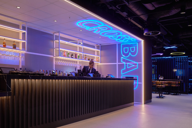 A modern bar with neon blue lights displaying 'Cross Bar'. A bartender stands behind the counter, surrounded by a selection of bottles on illuminated shelves.