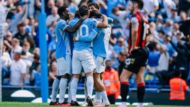 A group of soccer players in blue jerseys are hugging each other on the field, celebrating a goal, with a player in a red and black jersey walking past.