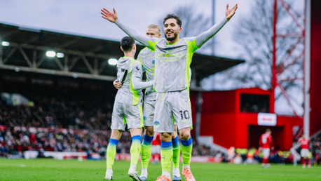 Three Manchester City players celebrating a goal on the field, with the number 10 player raising arms. They are wearing the team's kit with Etihad Airways sponsorship visible.