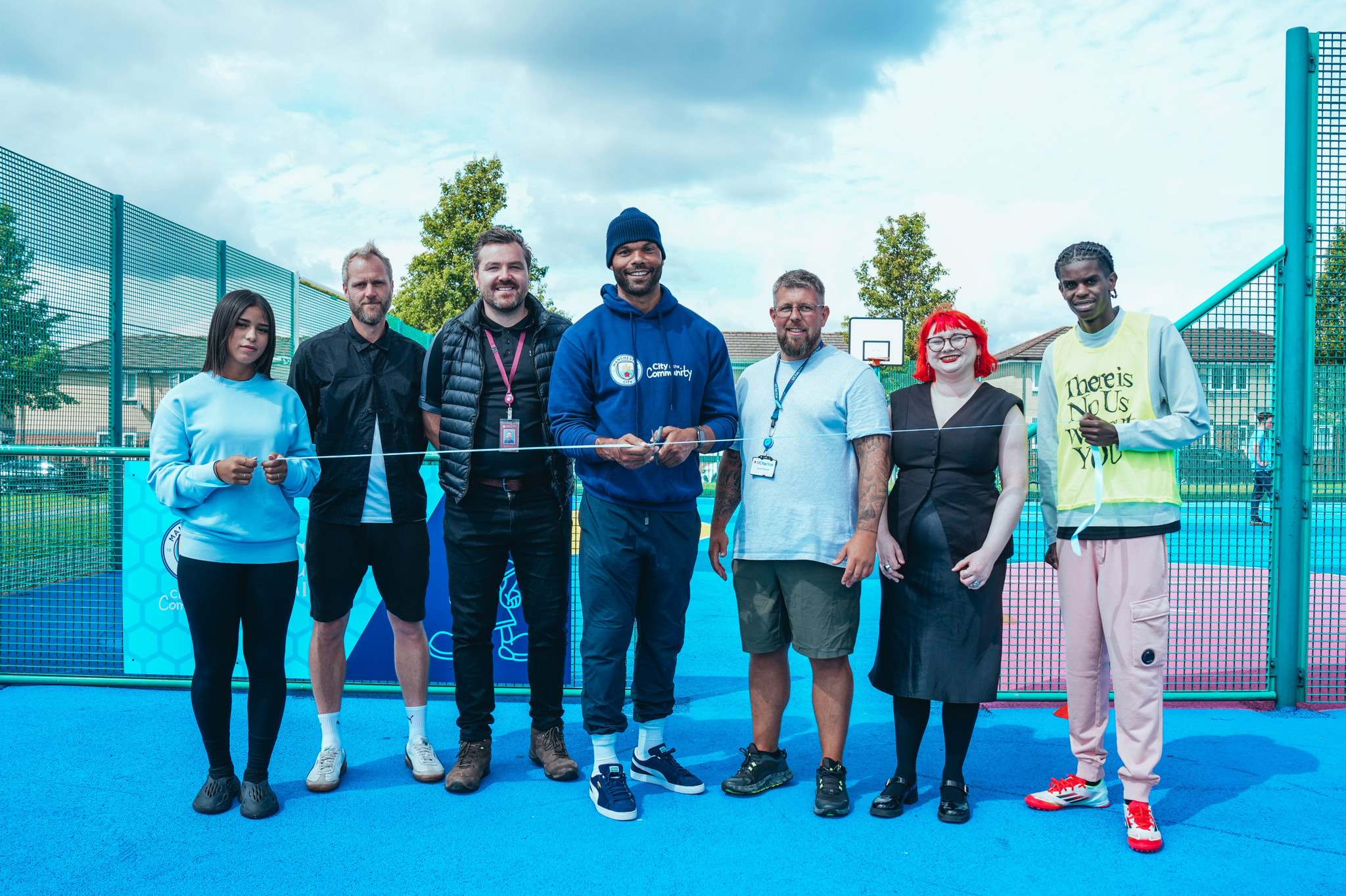 Group of people standing on a blue outdoor sports court participating in a ribbon-cutting ceremony. The middle person is holding scissors and wearing a blue 'City in the Community' sweatshirt.