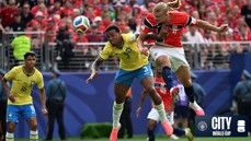 A soccer match featuring players from Brazil and Norway competing for a header. The background shows a crowded stadium with spectators wearing red attire.