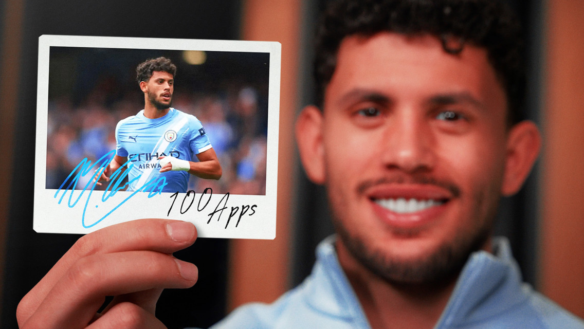 A person holds a signed photo featuring a Manchester City player in the club’s kit, annotated with '100 Apps'.