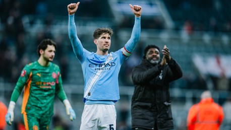 Manchester City player in a light blue jersey with the Etihad Airways logo raises his arms in celebration on a football pitch. Teammates applaud around him.