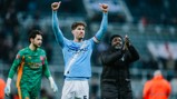 Manchester City player in a light blue jersey with the Etihad Airways logo raises his arms in celebration on a football pitch. Teammates applaud around him.