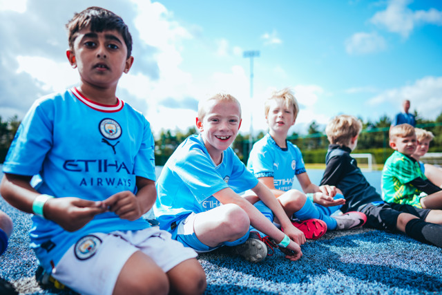 Group of young boys wearing Manchester City jerseys sitting on the ground with their faces blurred.