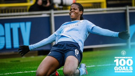 Manchester City player in a light blue kit performs a goal celebration sliding on her knees with arms outstretched. Text indicates '100 Goals Bunny Shaw 2021/22 - Present'.