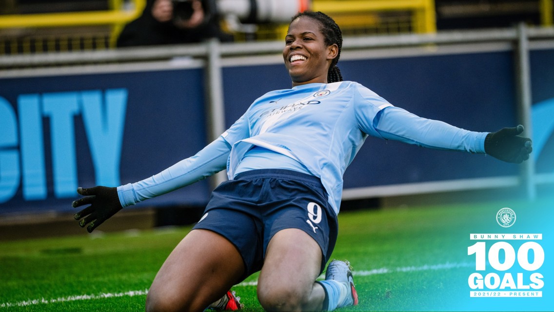 Manchester City player in a light blue kit performs a goal celebration sliding on her knees with arms outstretched. Text indicates '100 Goals Bunny Shaw 2021/22 - Present'.