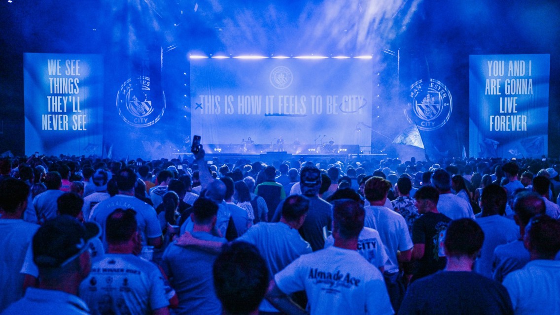 A large crowd of Manchester City fans in a concert-like setting with banners displaying celebratory messages like 'This is how it feels to be City'.