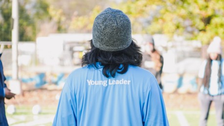 A person wearing a blue shirt with 'Young Leader' written on the back, standing on a football field, blurred in focus with activities happening in the background.