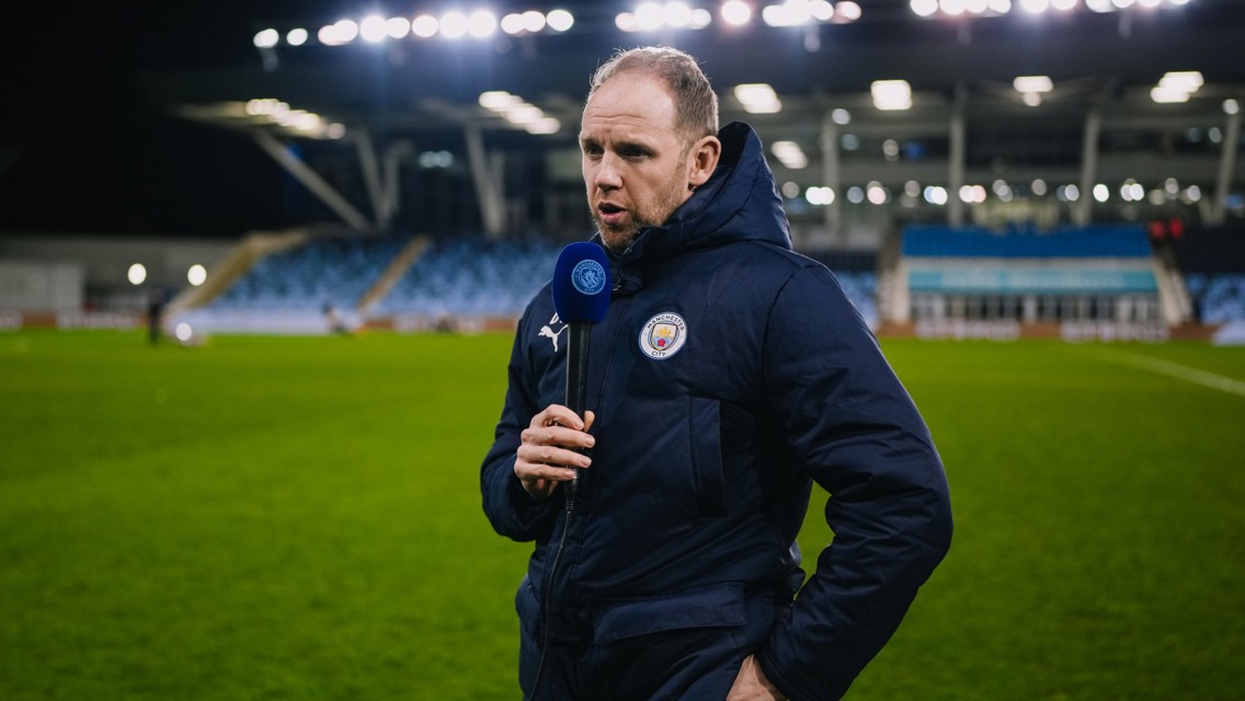 Person holding a microphone with Manchester City logo at a stadium, standing on grass with empty stands and bright floodlights.