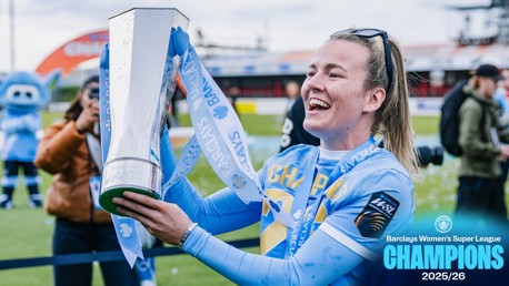 A person in Manchester City kit holds the Barclays Women's Super League trophy for the 2025/26 season. A mascot is visible in the background.