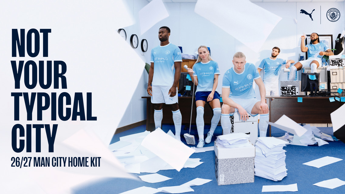 Five people in Manchester City 26/27 home kit in an office setting, with papers flying and a large text reading 'Not Your Typical City'.