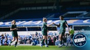 Manchester City women's team players on a soccer field celebrating a goal with a handshake, wearing green jerseys with Etihad sponsor, in a stadium with blue seating.