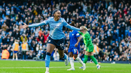 A Manchester City player celebrating a goal in front of a crowd at a stadium. The player is wearing a light blue kit, and the background shows fans cheering.