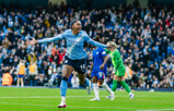 A Manchester City player celebrating a goal in front of a crowd at a stadium. The player is wearing a light blue kit, and the background shows fans cheering.