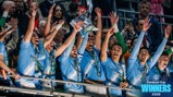 Football players in light blue jerseys celebrate lifting a silver trophy on a podium with crowd in background. The text 'Carabao Cup Winners 2026' is visible.