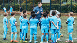 A group of young boys in blue football uniforms gather around a coach on a football field. The coach appears to be giving instructions.