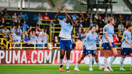 A women's soccer team in light blue uniforms celebrates on the field in front of a cheering crowd, with one player raising her arms.