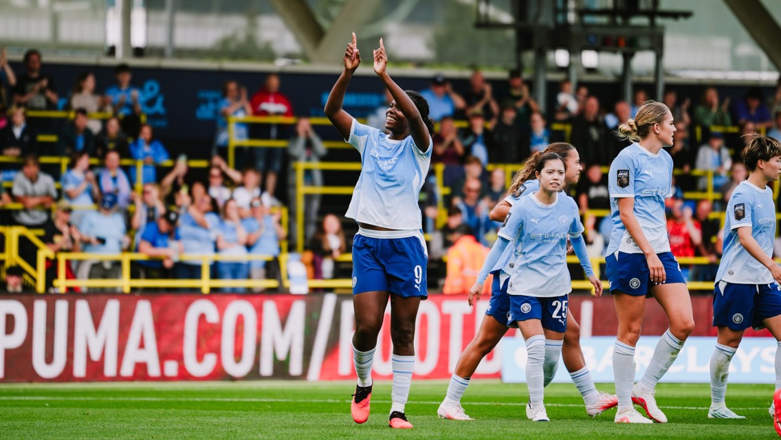 A women's soccer team in light blue uniforms celebrates on the field in front of a cheering crowd, with one player raising her arms.