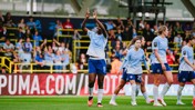 A women's soccer team in light blue uniforms celebrates on the field in front of a cheering crowd, with one player raising her arms.