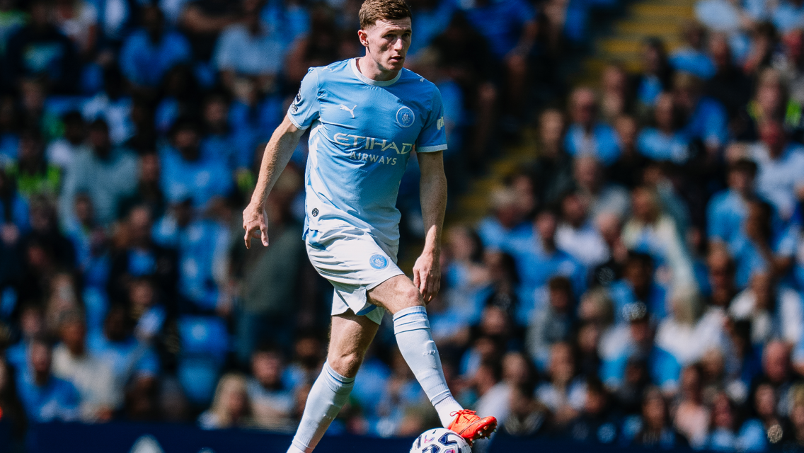 A Manchester City player in focus during a match, wearing the team's light blue jersey and kicking a football on the field.