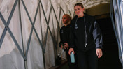 Two people wearing black jackets walking through a tunnel-like structure, with one holding a bottle and earbuds.