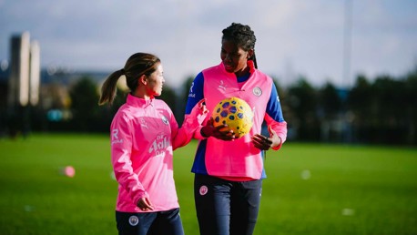 Two soccer players in pink training kits on a grass field, one holding a ball with a Manchester City logo visible.