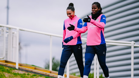 Two football players in Manchester City training kits walking on a path, wearing gloves and discussing.