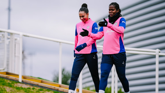 Two football players in Manchester City training kits walking on a path, wearing gloves and discussing.