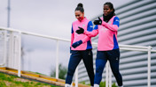 Two football players in Manchester City training kits walking on a path, wearing gloves and discussing.