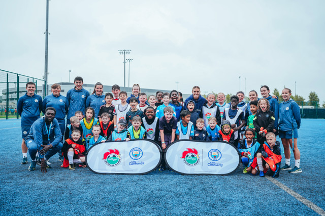 Group photo of Manchester City community event participants including children and coaches on a blue pitch.