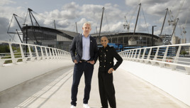 Two people standing on a bridge with the Etihad Stadium in the background, one in a casual blazer and jeans, the other in a black stylish ensemble.