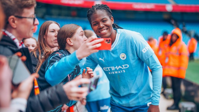 A Manchester City football player taking a selfie with fans at the Etihad Stadium. Faces of fans and the player are blurred.